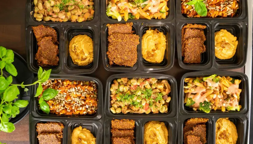 Vorbereitete Mahlzeiten in schwarzen Behältern: Pasta mit Brokkoli, Linsen mit Sonnenblumenkernen und Bohnen-Eintopf. Dazu Flachbrot und Hummus – perfekt für low carb high protein.