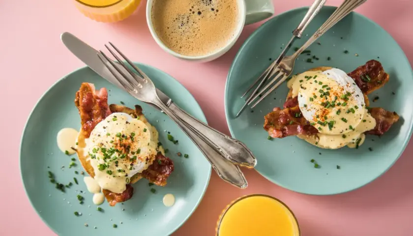 Zwei Teller mit Eggs Benedict, dazu Kaffee und Orangensaft auf rosa Untergrund.
