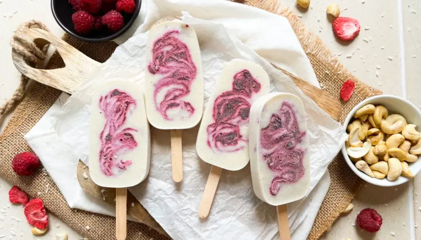 Vier selbstgemachte Eis am Stiel mit Himbeermuster, umgeben von frischen Himbeeren, Cashews und gefriergetrockneten Erdbeeren.