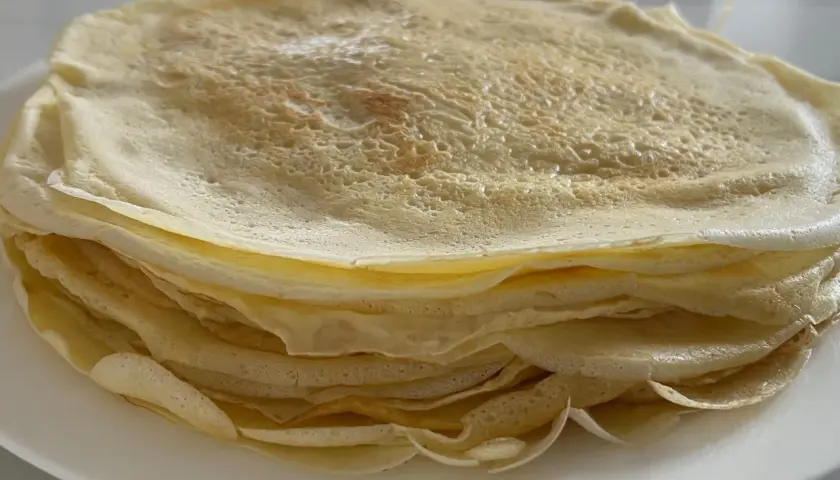 Ein Stapel goldbrauner Crêpes, frisch zubereitet aus dem Teig, liegt auf einem weißen Teller.