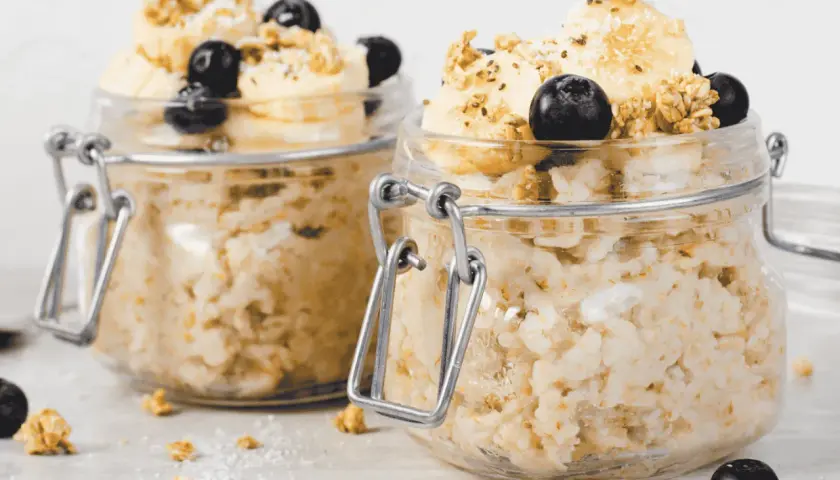 Zwei Gläser mit Overnight Oats, garniert mit Bananenscheiben, Blaubeeren und Granola. Perfekt für ein schnelles Frühstück.