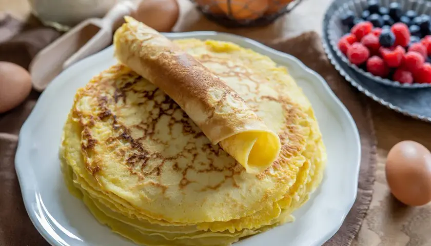 Ein Stapel goldbrauner Pfannkuchen, einer davon aufgerollt, wartet auf ein leckeres Pfannkuchen Rezept. Beeren und Eier im Hintergrund.