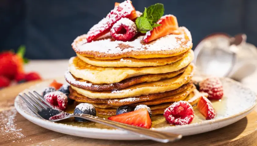 Ein Stapel fluffiger amerikanischer Pancakes, bestäubt mit Puderzucker und garniert mit frischen Beeren und Minze. Ein köstliches american pancakes rezept für den perfekten Brunch.