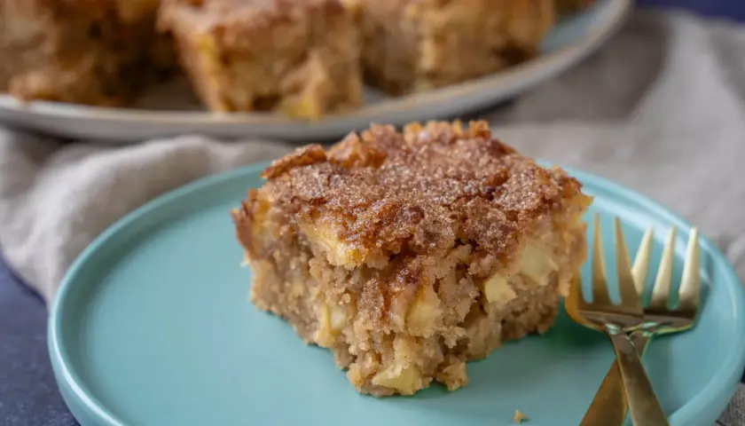 Ein Stück saftiger Apfel Zimt Kuchen mit knuspriger Kruste auf einem türkisfarbenen Teller, daneben zwei goldene Gabeln.