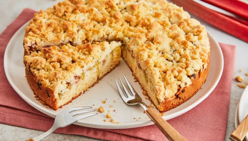 Ein saftiger Rhabarberkuchen mit Streuseln, bereit zum Genießen. Die roten Rhabarberstangen im Hintergrund versprechen Frische.