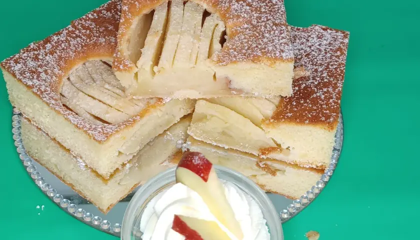 Saftiger Apfelkuchen vom Blech, mit Puderzucker bestäubt und auf einem Spiegel serviert. Ein Stück mit Sahne und Apfel garniert.