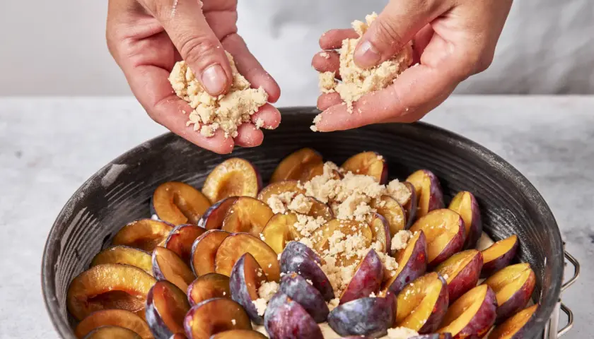 Hände streuen Streusel über saftige Pflaumen in einer Springform. Ein köstlicher saftiger Pflaumenkuchen entsteht.
