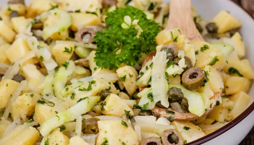 Ein köstlicher Kartoffelsalat ohne Mayo mit Gurken, Oliven und Kapern, garniert mit Petersilie.