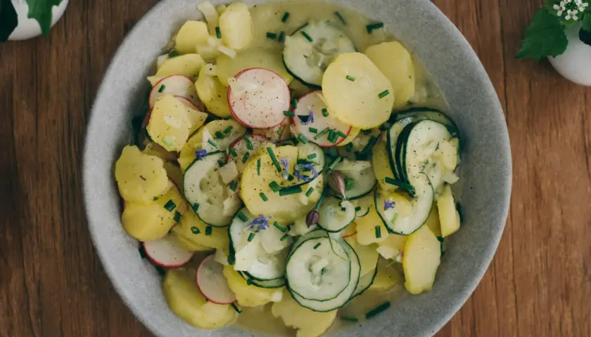 Bayrischer Kartoffelsalat mit Speck und Zwiebeln, garniert mit Radieschen, Gurken und Schnittlauch. Ein deftiger Genuss!