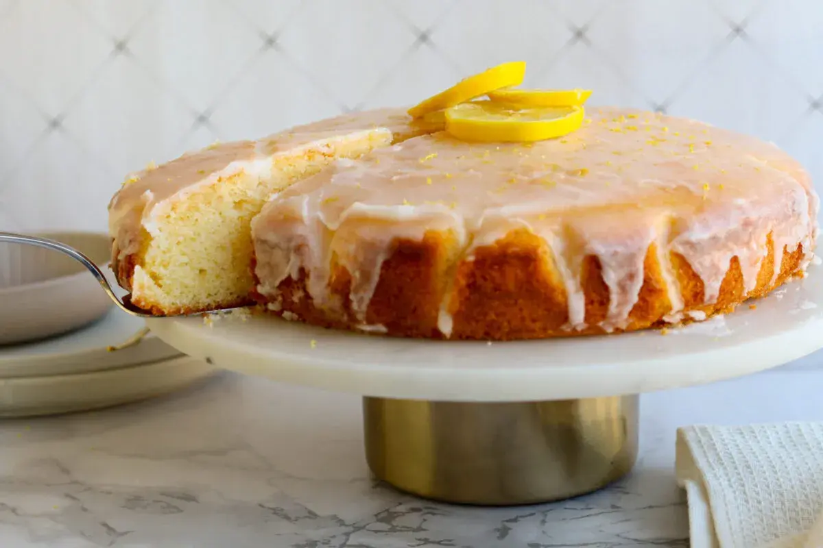 Ein saftiger Zitronenkuchen mit Zuckerguss und Zitronenscheiben, der beste Zitronenkuchen der Welt, wird auf einer Tortenplatte präsentiert.
