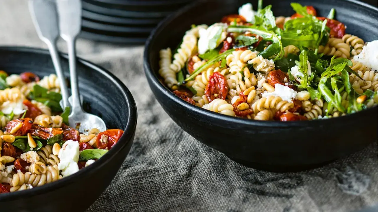 Zwei Schalen mit Nudelsalat mit getrockneten Tomaten, Pinienkernen und Rucola. Ein Löffel liegt in der linken Schale.