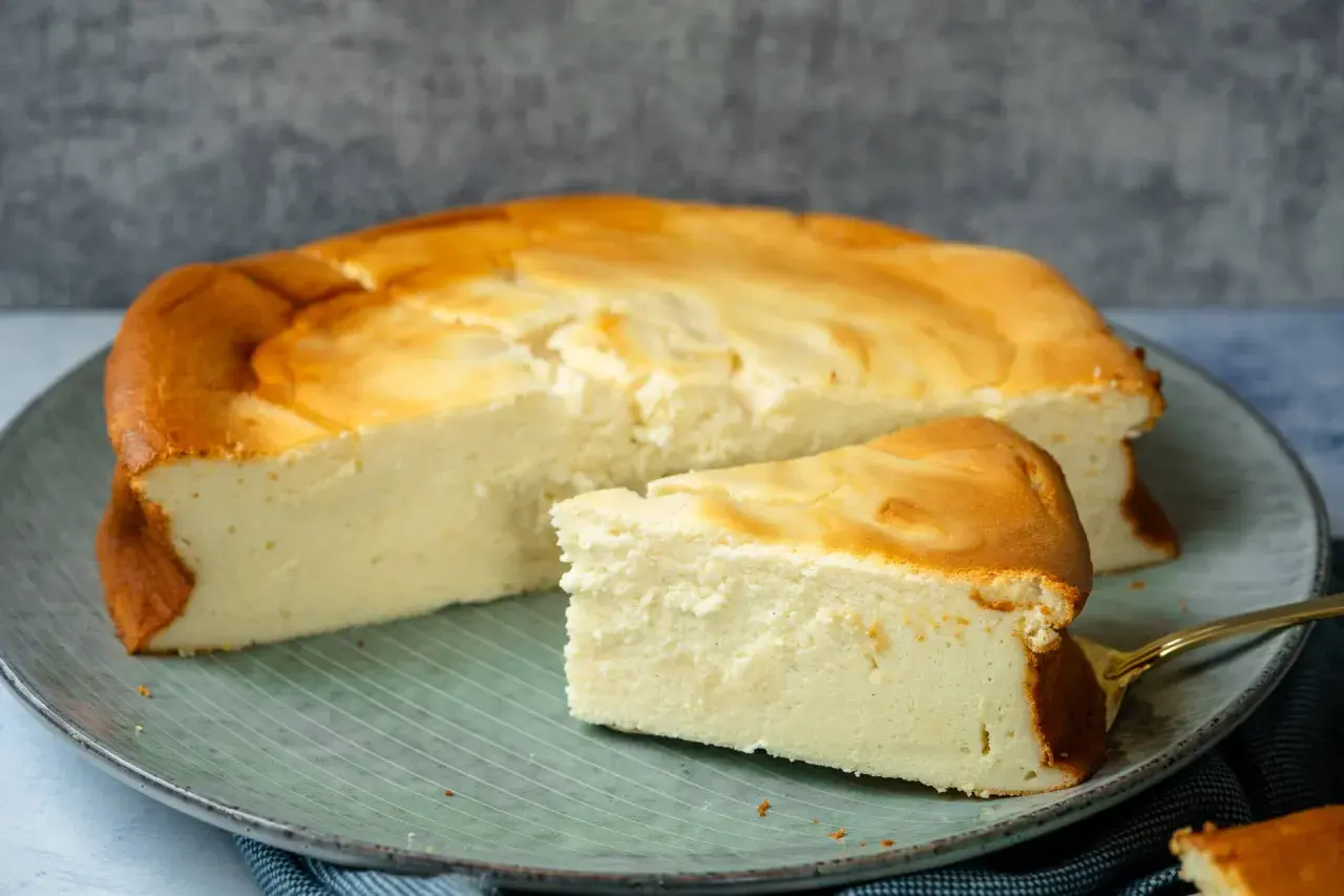 Ein saftiger Quarkkuchen ohne Boden, goldbraun gebacken, liegt auf einem Teller. Ein Stück ist bereits angeschnitten.