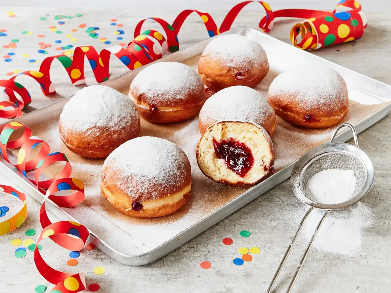 Berliner Rezept Original: Sieben luftige Krapfen mit Marmeladenfüllung, bestäubt mit Puderzucker, auf einem Blech, umgeben von bunten Party-Girlanden und Konfetti.