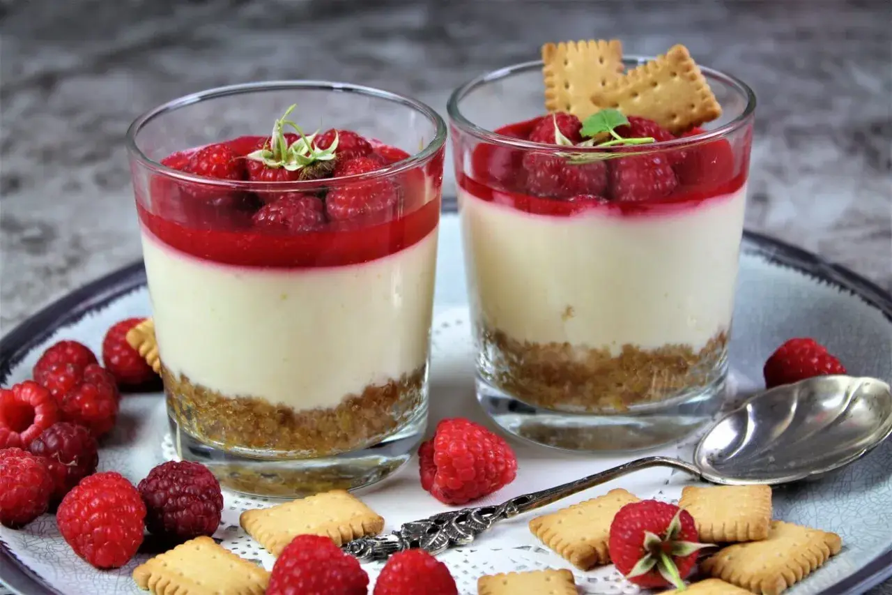 Zwei Gläser mit Himbeer-Dessert: eine Schicht Keksbrösel, cremige Masse und frische Himbeeren mit Himbeersauce.