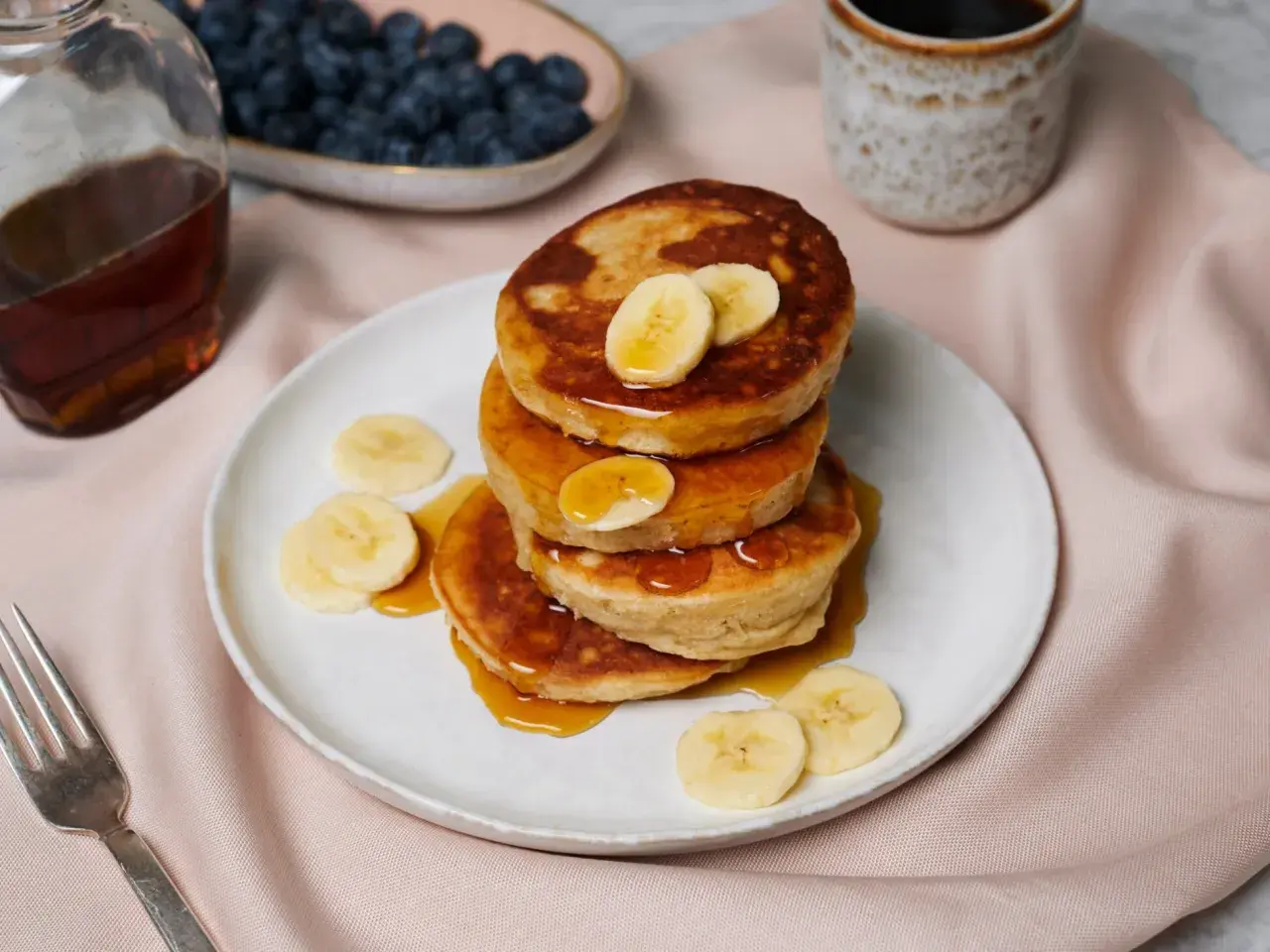 Ein Stapel goldbrauner Bananen-Pancakes, beträufelt mit Sirup und garniert mit Bananenscheiben. Daneben Blaubeeren, Ahornsirup und Kaffee.