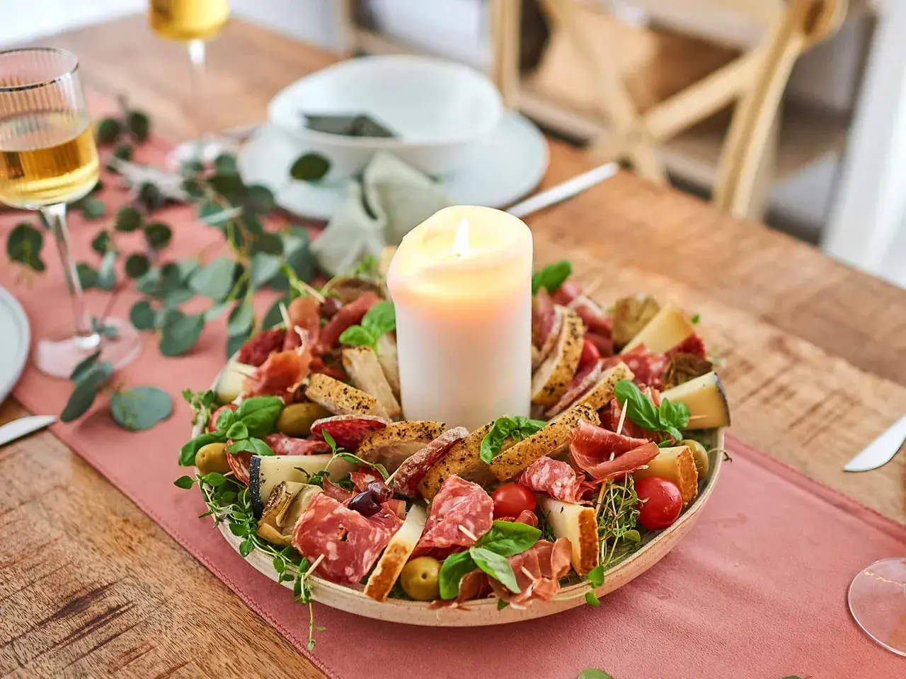 Eine festliche Platte mit Wurst, Käse, Brot und Oliven, die zum Antipasti selber machen einlädt. Ein Kerzenlicht sorgt für Gemütlichkeit.