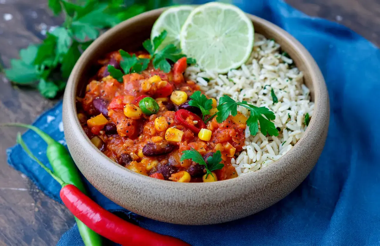 Herzhaftes Chili sin Carne mit Reis, garniert mit frischer Petersilie und Limettenspalten. Rote und grüne Chilis liegen daneben.