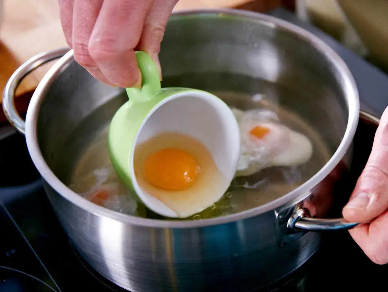 Ein rohes Ei wird aus einer grünen Tasse in kochendes Wasser gegossen. Ein Schritt für ein perfektes verlorene Eier Rezept.