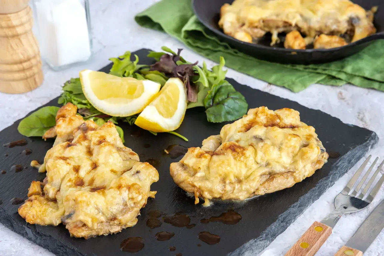 Zwei Portionen überbackenes Steak au four mit geschmolzenem Käse, serviert mit Salat und Zitronenspalten auf einem schwarzen Schiefer.