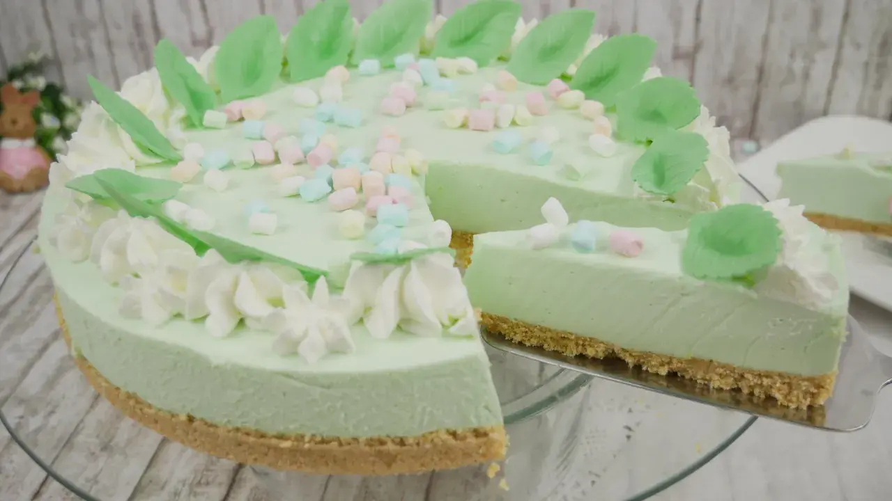 Eine helle, cremige Philadelphia Torte mit Waldmeistergeschmack, dekoriert mit Marshmallows und grünen Blättern. Ein Stück wird gerade serviert.