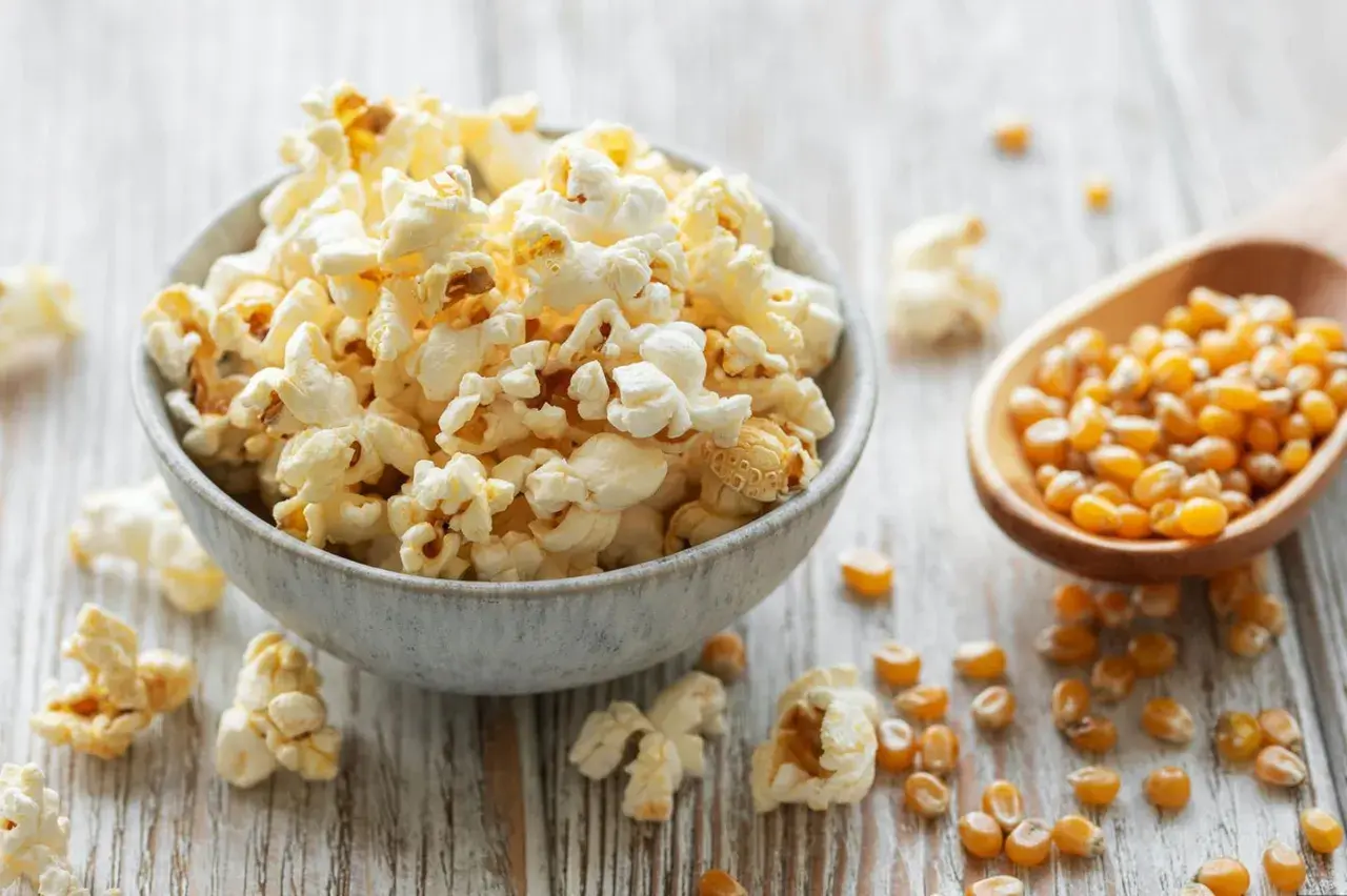 Schüssel mit frisch gemachtem Popcorn, daneben Maiskörner in einem Holzlöffel. So einfach ist Popcorn selber machen!