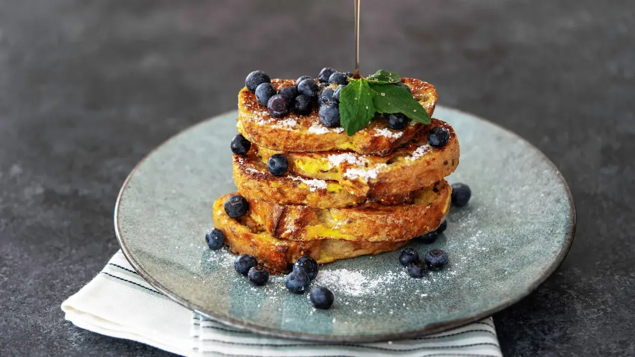 Ein Stapel goldbrauner French Toast, garniert mit Blaubeeren und Puderzucker. Ein dünner Strahl Sirup fließt darüber.