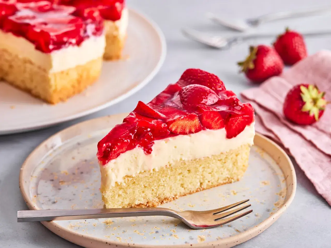 Ein saftiges Stück Erdbeer Schmand Kuchen mit frischen Erdbeeren und Tortenguss auf einem Teller, daneben frische Erdbeeren und eine Gabel.