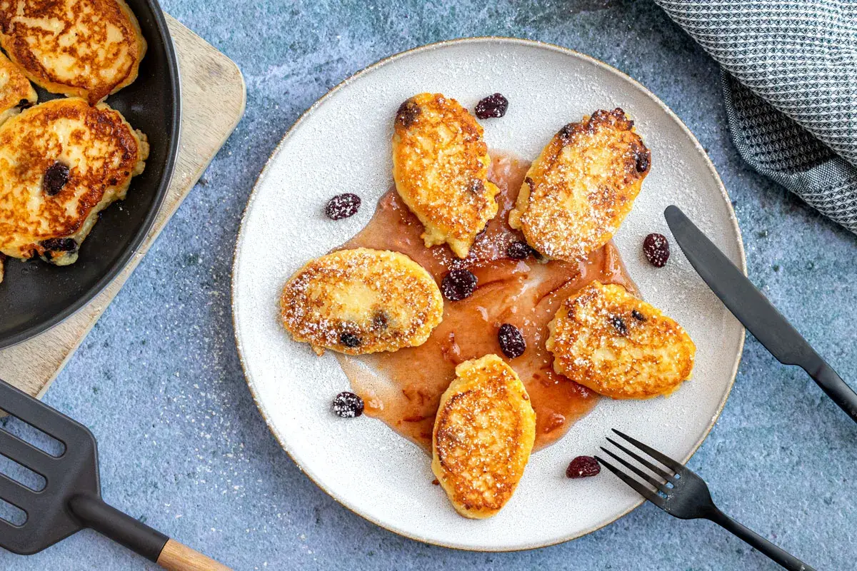 Goldbraune Quarkkeulchen ohne Kartoffeln, mit Puderzucker bestäubt und mit Rosinen garniert, serviert mit fruchtiger Soße.