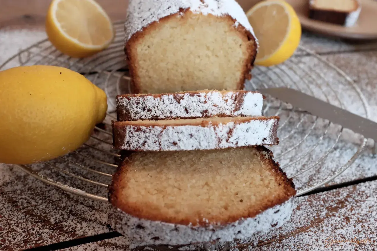 Zitronenkuchen wie bei Oma, mit Puderzucker bestäubt, neben frischen Zitronen auf einem Gitterrost.