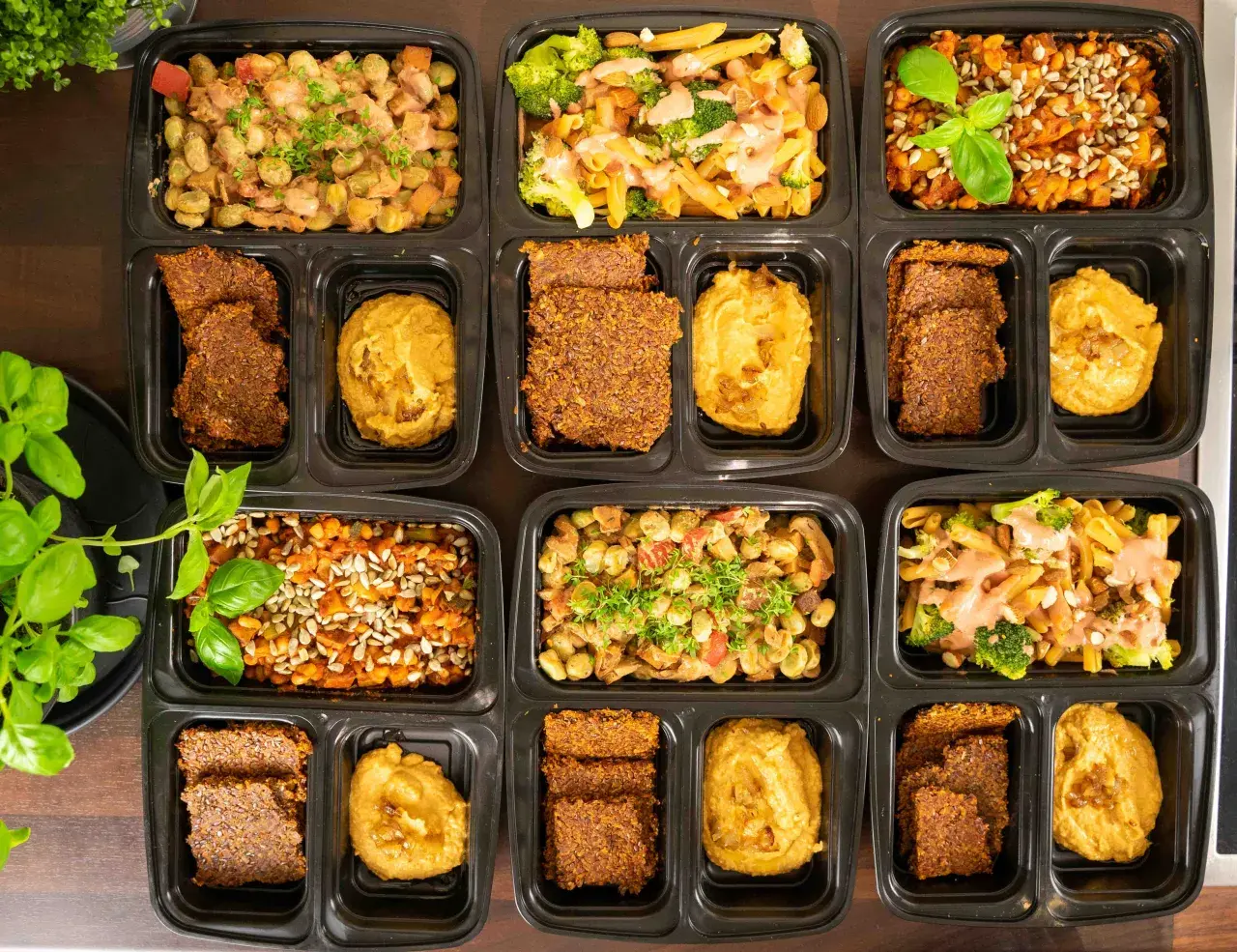 Vorbereitete Mahlzeiten in schwarzen Behältern: Pasta mit Brokkoli, Linsen mit Sonnenblumenkernen und Bohnen-Eintopf. Dazu Flachbrot und Hummus – perfekt für low carb high protein.