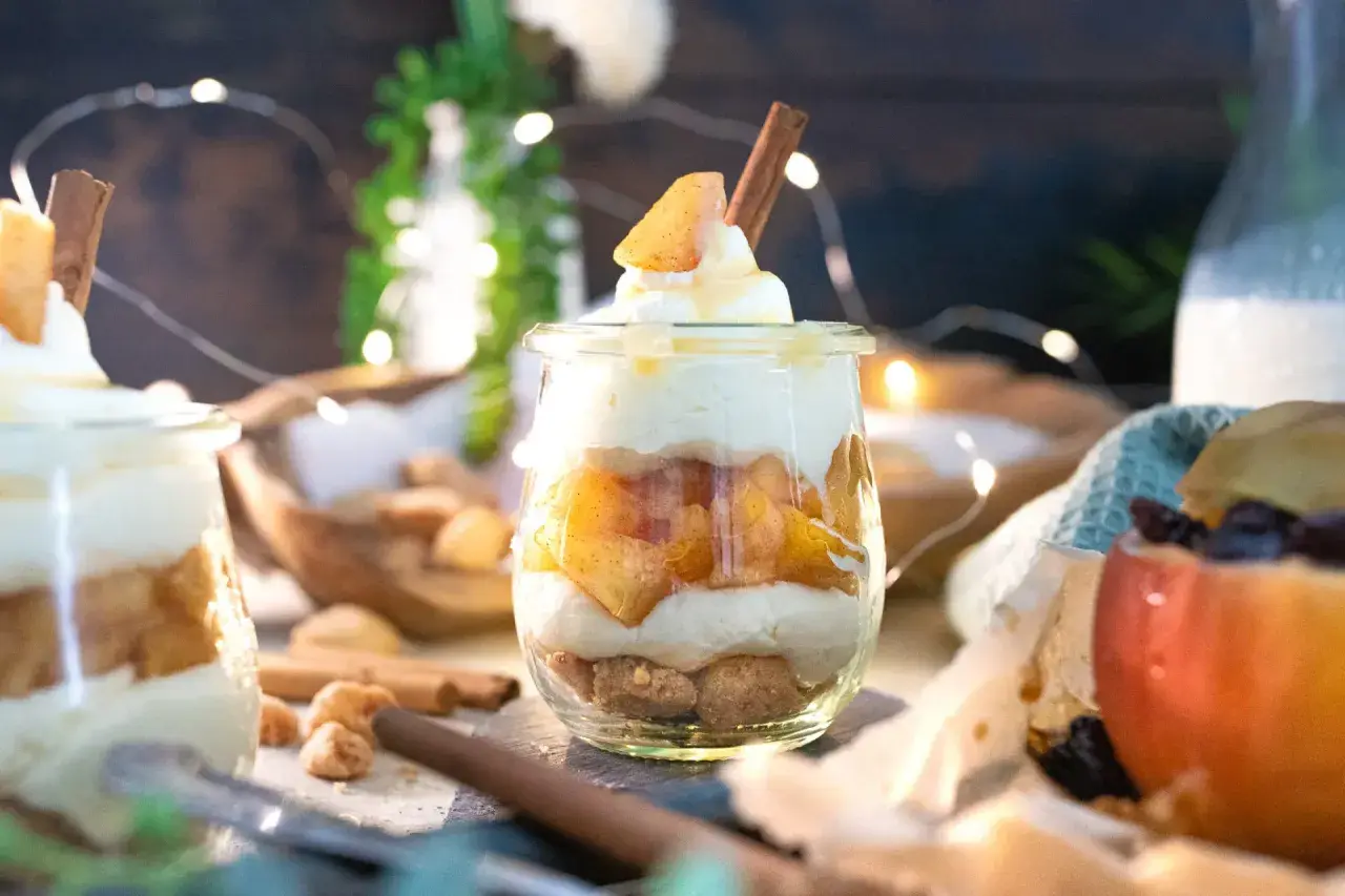 Leckeres Apfel-Crumble-Dessert im Glas mit Zimtstange und Sahne. Ein süßer Genuss, der zum Naschen einlädt.