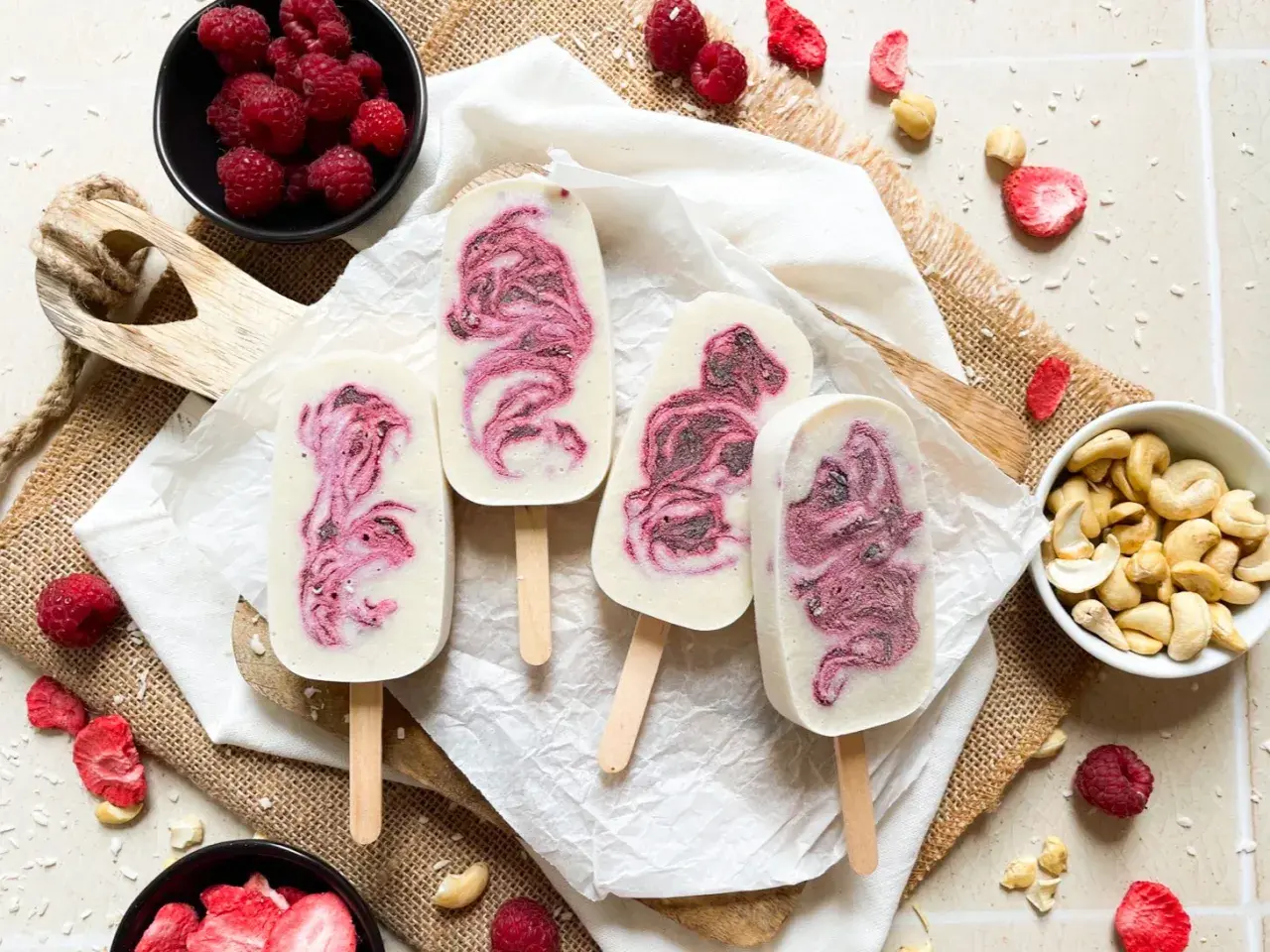 Vier selbstgemachte Eis am Stiel mit Himbeermuster, umgeben von frischen Himbeeren, Cashews und gefriergetrockneten Erdbeeren.