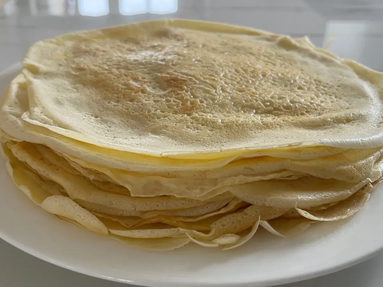 Ein Stapel goldbrauner Crêpes, frisch zubereitet aus dem Teig, liegt auf einem weißen Teller.