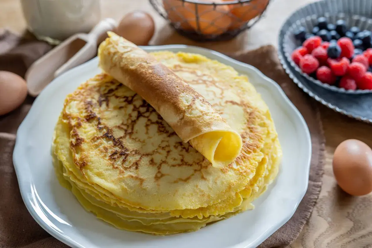 Ein Stapel goldbrauner Pfannkuchen, einer davon aufgerollt, wartet auf ein leckeres Pfannkuchen Rezept. Beeren und Eier im Hintergrund.