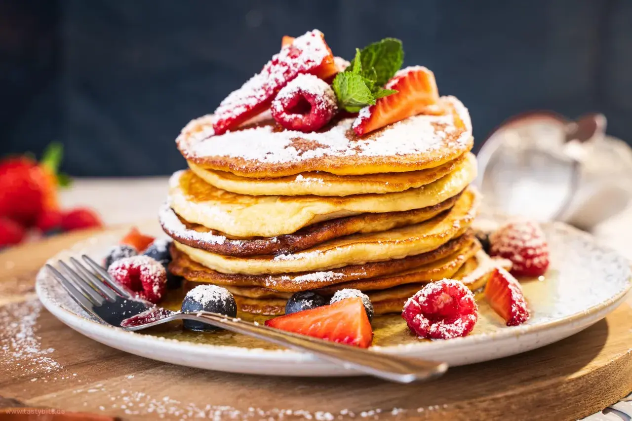 Ein Stapel fluffiger amerikanischer Pancakes, bestäubt mit Puderzucker und garniert mit frischen Beeren und Minze. Ein köstliches american pancakes rezept für den perfekten Brunch.