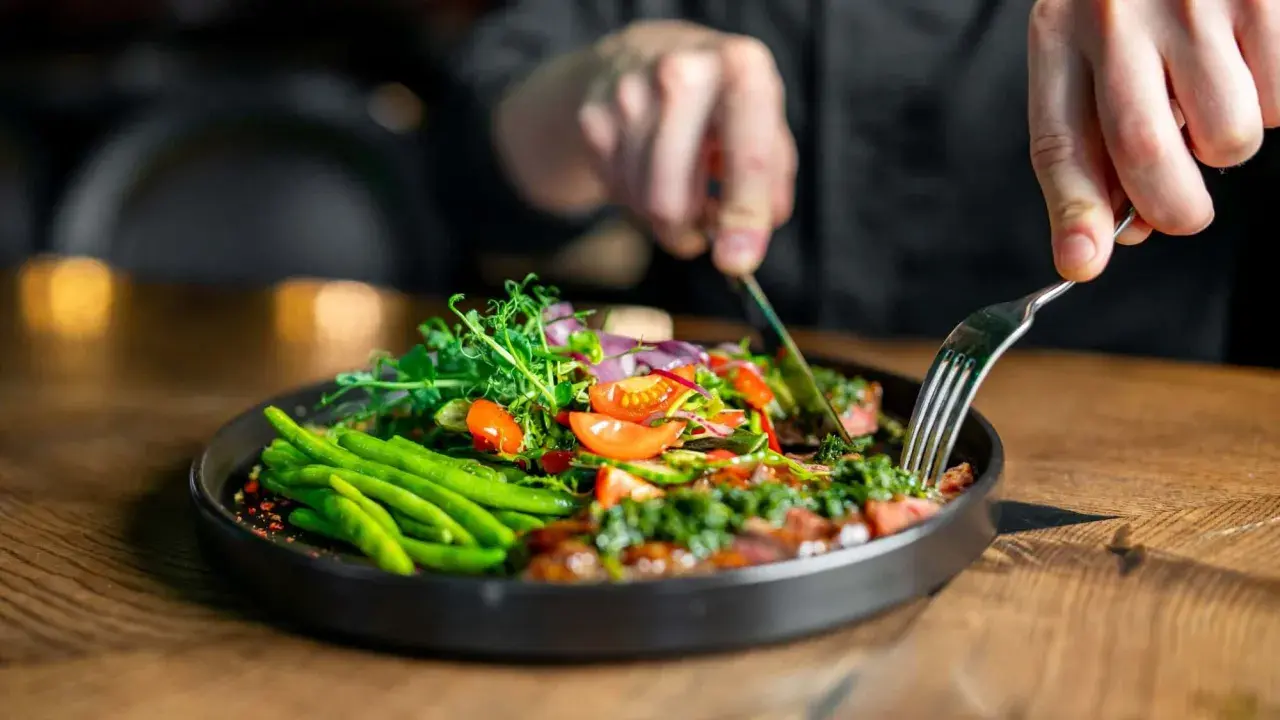 Ein Teller mit frischem Gemüse, wie grüne Bohnen und Tomaten, inspiriert von clean eating rezepten. Eine Person schneidet das Essen mit Messer und Gabel.