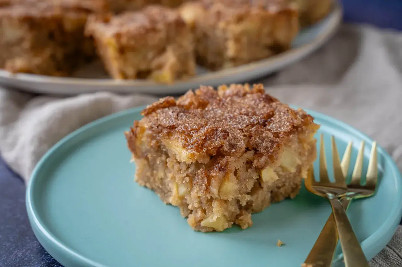 Ein Stück saftiger Apfel Zimt Kuchen mit knuspriger Kruste auf einem türkisfarbenen Teller, daneben zwei goldene Gabeln.