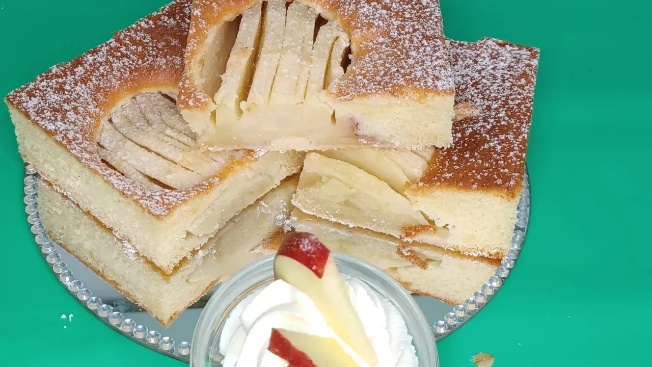 Saftiger Apfelkuchen vom Blech, mit Puderzucker bestäubt und auf einem Spiegel serviert. Ein Stück mit Sahne und Apfel garniert.