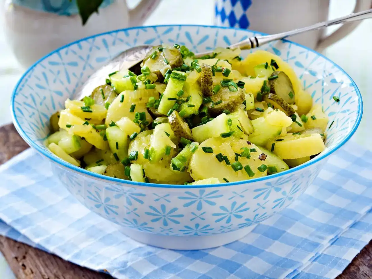 Schüssel mit Kartoffelsalat ohne Mayo, garniert mit Gurken und Schnittlauch. Ein Löffel liegt darin.