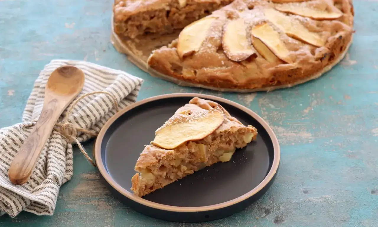 Ein Stück saftiger Apfelkuchen ohne Zucker, mit Apfelscheiben belegt und mit Puderzucker bestäubt, auf einem Teller neben einem Holzlöffel und einer Serviette.
