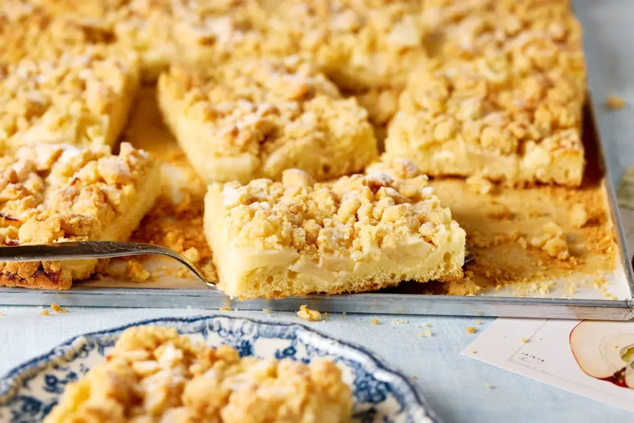 Saftiger apfelkuchen mit vanillepudding und Streuseln, frisch aus dem Ofen. Ein St&uuml;ck wird gerade herausgehoben.
