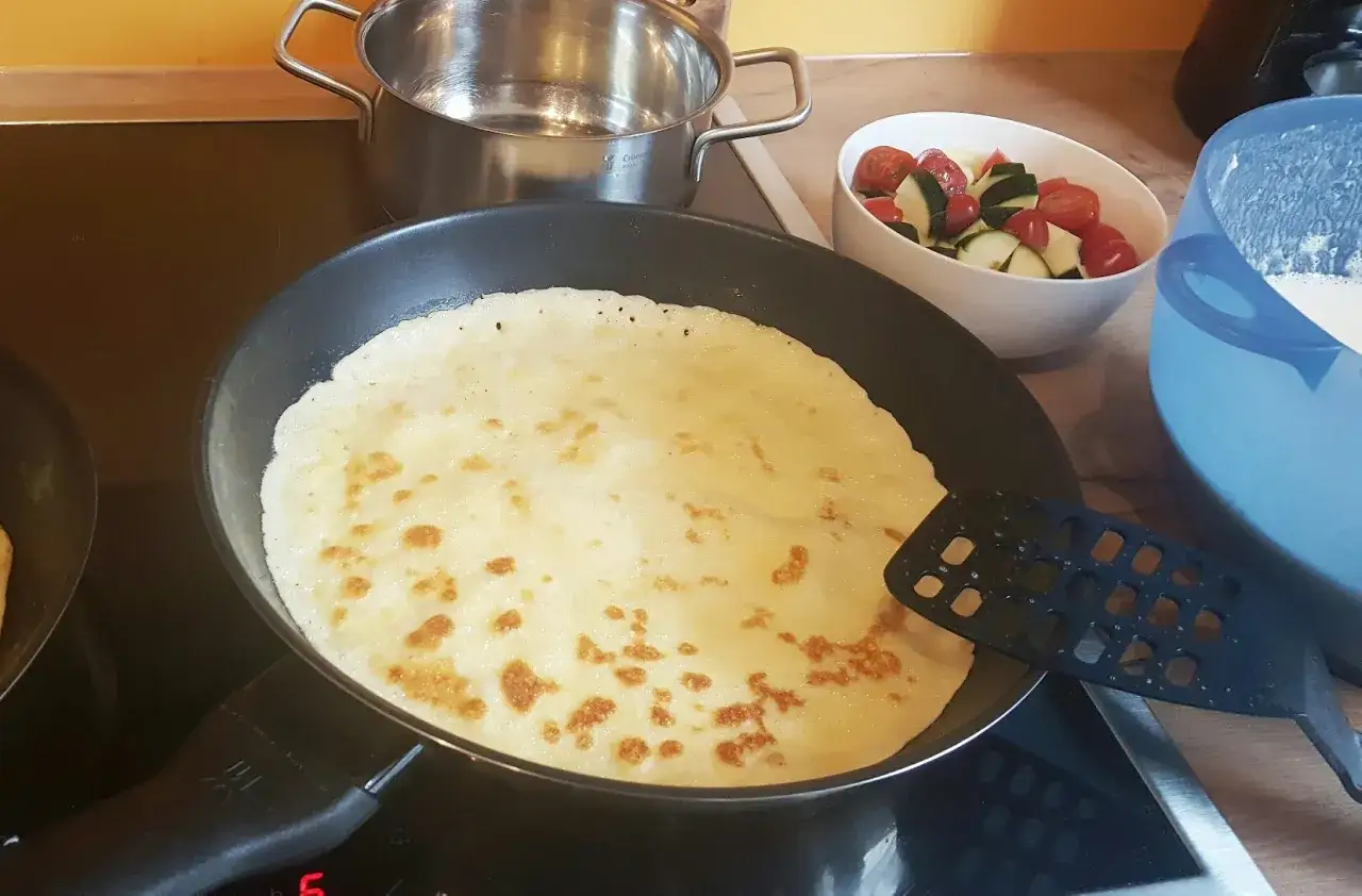 Ein Pfannkuchen wird in der Pfanne goldbraun gebacken. Daneben stehen Zutaten f&uuml;r ein Pfannkuchen rezept: ein Topf, eine Sch&uuml;ssel mit Tomaten und Gurken und eine Sch&uuml;ssel mit Teig.