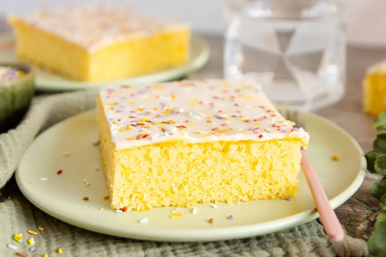Ein St&uuml;ck gelber Blechkuchen mit wei&szlig;er Glasur und bunten Streuseln. Perfekt f&uuml;r ein schnelles Tassenkuchen Rezept vom Blech.
