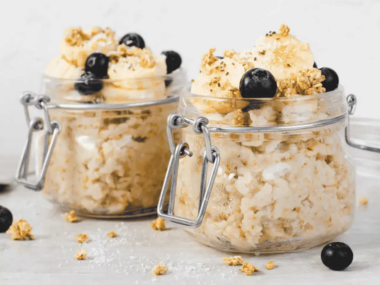Zwei Gläser mit Overnight Oats, garniert mit Bananenscheiben, Blaubeeren und Granola. Perfekt für ein schnelles Frühstück.