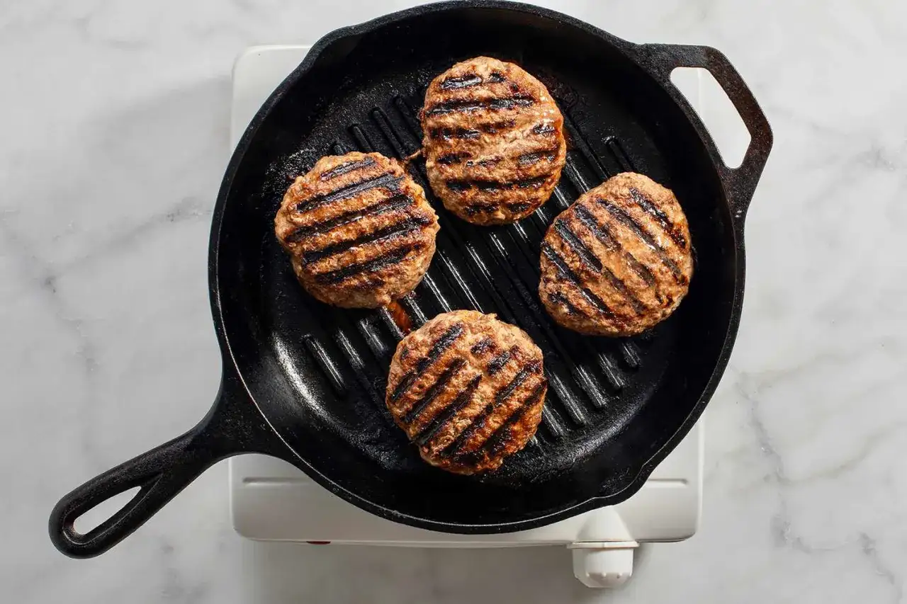 Vier saftige Burger-Patties brutzeln in einer Gusseisenpfanne und entwickeln köstliche Grillstreifen.
