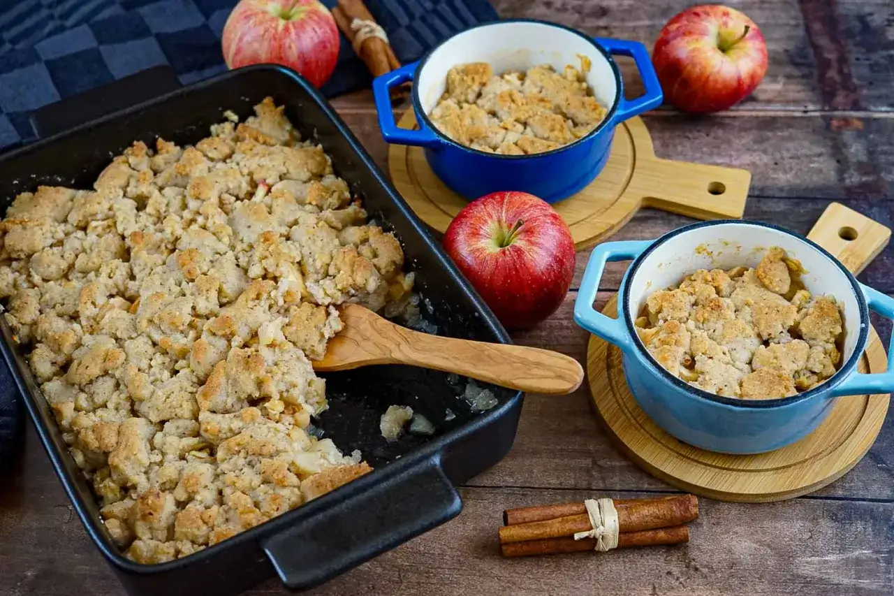 Ein frisch gebackener Apfel Crumble in einer schwarzen Auflaufform und zwei blauen Förmchen. Daneben liegen Äpfel und Zimtstangen.