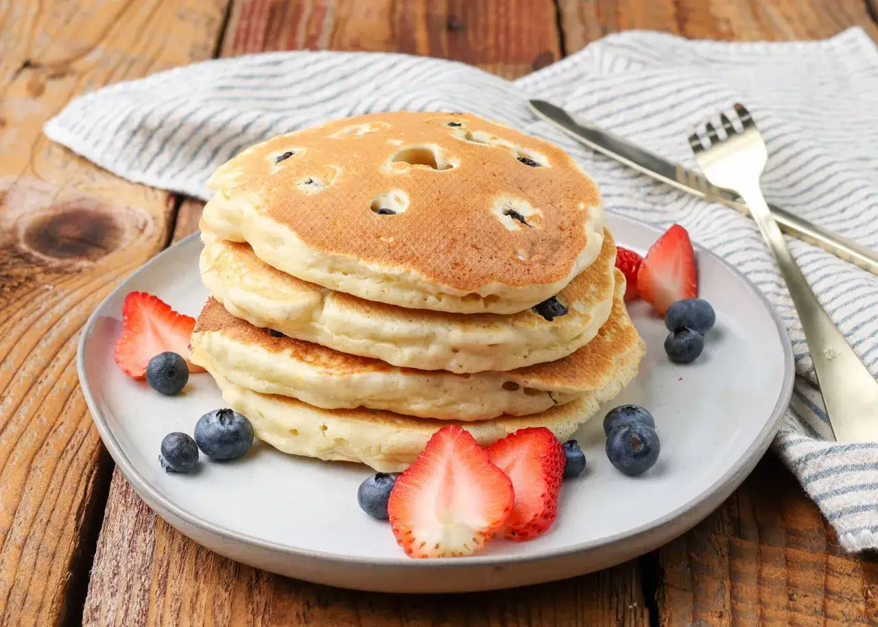 Ein Stapel fluffiger amerikanischer Pancakes mit Blaubeeren, garniert mit Erdbeeren und Blaubeeren. Perfekt für ein amerikanisches Pancakes Rezept.