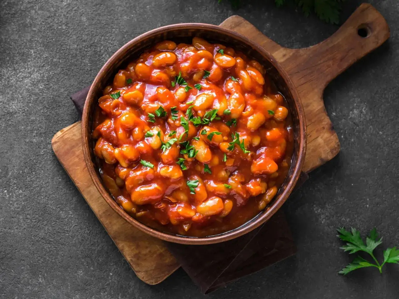Schmackhaftes baked beans rezept in einer Schüssel, garniert mit Petersilie, auf einem Holzbrett.