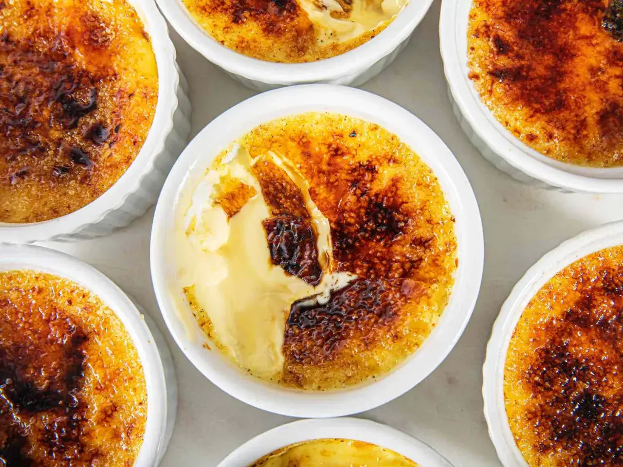 Mehrere cremige Crème brûlée in weißen Förmchen, mit karamellisierter Zuckerkruste, die beim Anbrechen aufbricht.