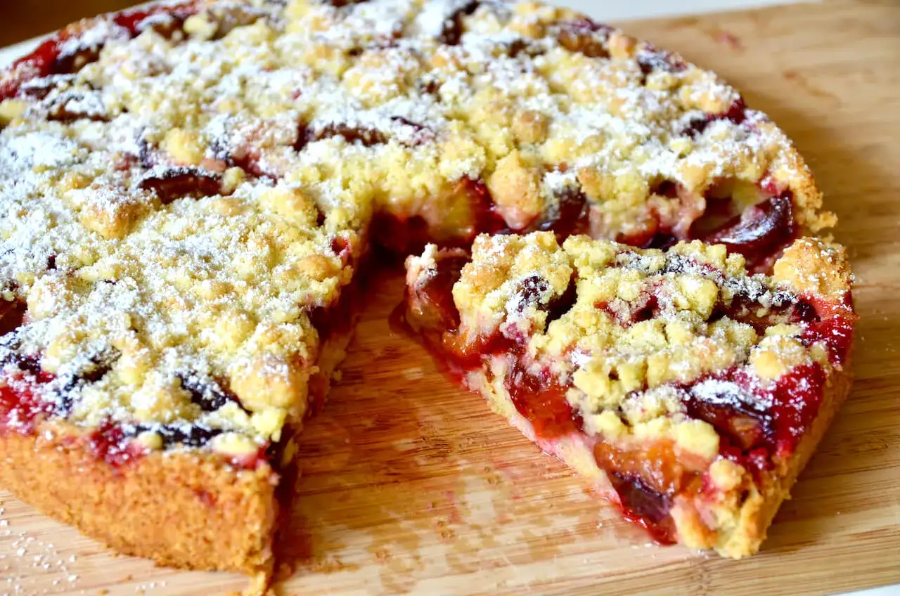 Saftiger Pflaumenkuchen in Springform, mit Streuseln und Puderzucker bestäubt. Ein Stück wurde herausgeschnitten, um die saftigen Pflaumen zu zeigen.